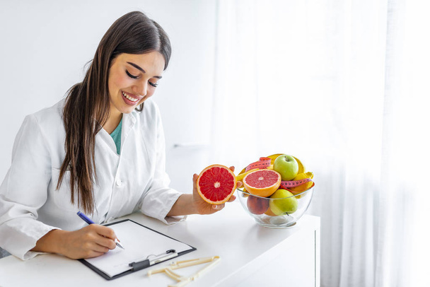 Usmívající se odbornice na výživu ve své kanceláři ukazuje zdravou zeleninu a ovoce, zdravotní a dietní koncepci. Žena nutriční s ovocem pracuje na svém stole.  - Fotografie, Obrázek