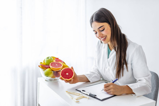 Portrait of young smiling female nutritionist in the consultation room. Nutritionist desk with healthy fruit, juice and measuring tape. Dietitian working on diet plan. - Foto, afbeelding