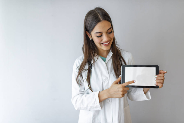 Portrait of smiling young doctor in white coat showing the screen of digital tablet in her hand. Doctor holding blank digital tablet isolated on white background with clipping path for the screen - 写真・画像
