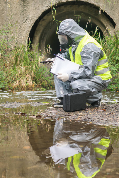 a specialist in a protective suit takes a photo of the place of alleged pollution of the reservoir using a mobile phone - Zdjęcie, obraz