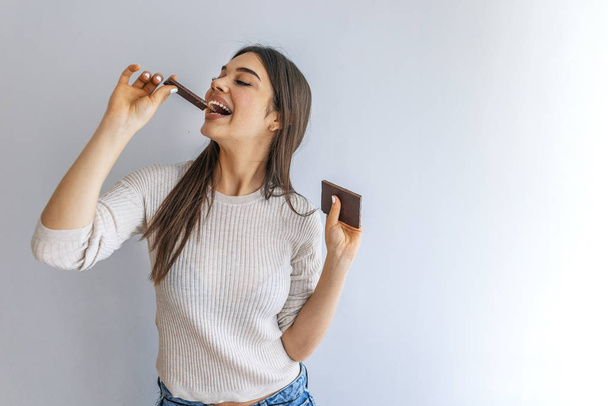Portrait of a happy young woman eating chocolate bar isolated over grey background. Happy young beautiful lady eating chocolate and smiling. Girl tasting sweet chocolate.  - Foto, Imagem