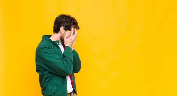 young crazy bearded man covering eyes with hands with a sad, frustrated look of despair, crying, side view against flat wall - Фото, изображение