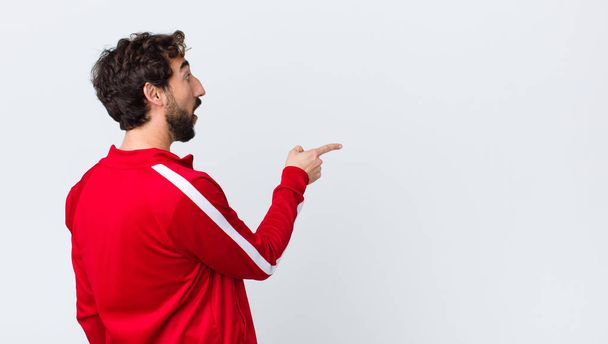young bearded man back view feeling shocked and surprised, pointing and looking upwards in awe with amazed, open-mouthed look against copy space wall - Fotó, kép