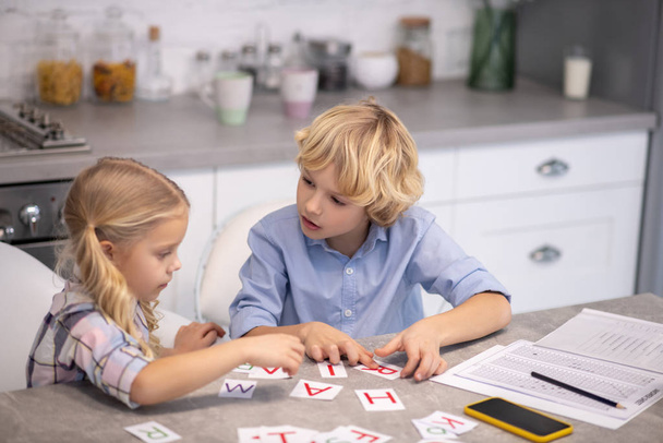Two blond kids putting cards with letters on the table and looking involved - Foto, Imagem