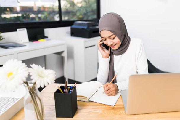 Beautiful Asian muslim woman working using laptop and mobile phone call in modern office. Small business company owner, startup entrepreneur, or working woman lifestyle concept - Photo, Image