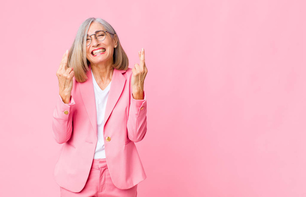 middle age cool woman smiling and anxiously crossing both fingers, feeling worried and wishing or hoping for good luck against flat wall - Fotoğraf, Görsel