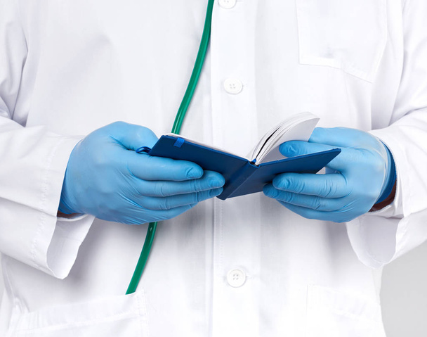 Arzt in weißem Mantel und blauen Handschuhen hält ein blaues Notizbuch in der Hand - Foto, Bild