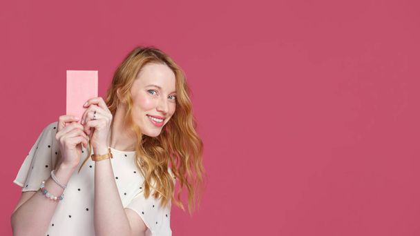 Cheerful blonde girl holds a pink notebook in her hands, looks at the camera and smiles, isolated on a pink background - Foto, Imagen