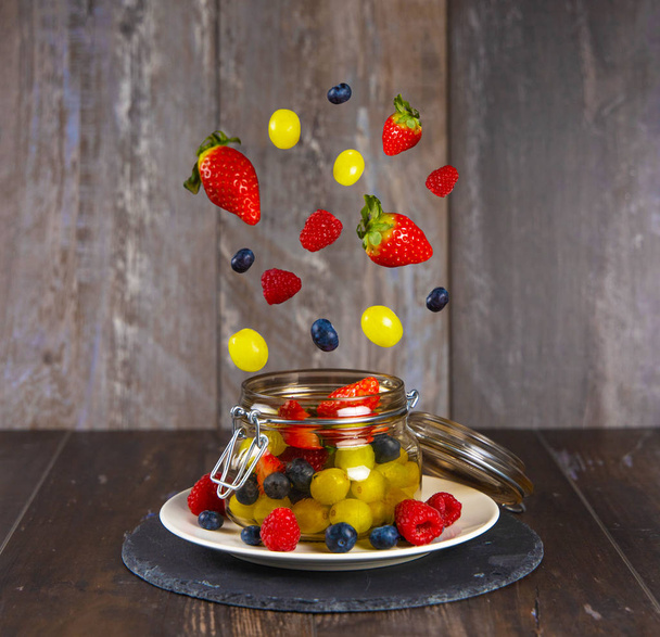 Owoce w ruchu, czerwone owoce spadające na szklany słoik. Winogrona, maliny, truskawki, jagody - Zdjęcie, obraz