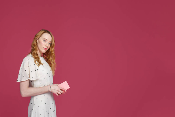 Cheerful blonde girl holds a pink notebook in her hands, looks at the camera and smiles, isolated on a pink background - Fotografie, Obrázek