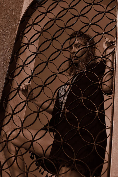 Vertikales Sepia-Porträt einer attraktiven jungen braunhaarigen Kaukasierin, die vor authentischem arabischen Hintergrund posiert, durch Gitter in die Kamera blickt und sich sicher fühlt. Mode, Stil, Schönheitskonzept. - Foto, Bild