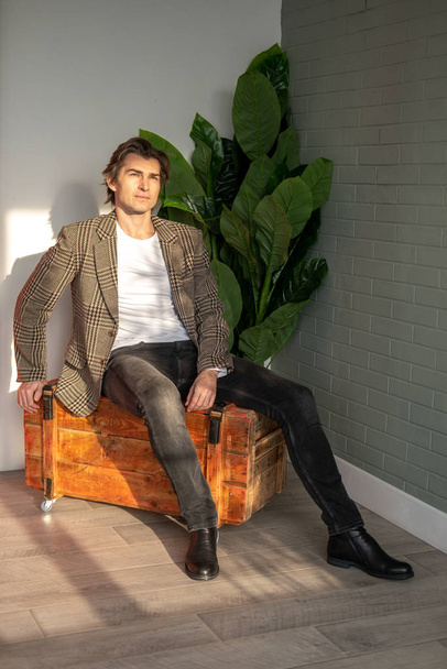 Tall handsome man sitting on the wooden box next to green plant and grey wall - Фото, изображение