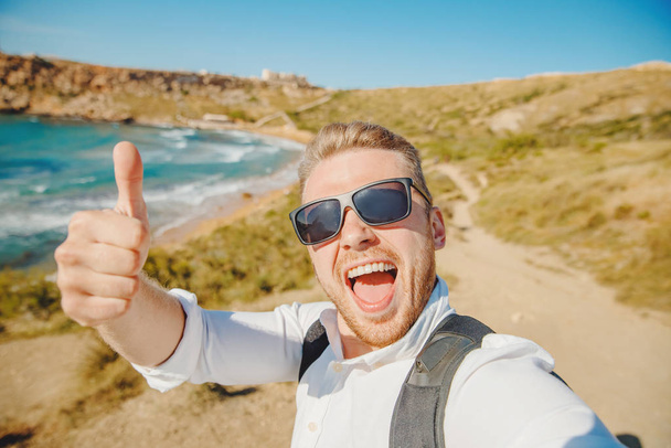 Male traveler makes selfie photo on background of blue sea, sand beach in sunglasses and with backpack. Travel concept. Gold bay Malta - Fotó, kép