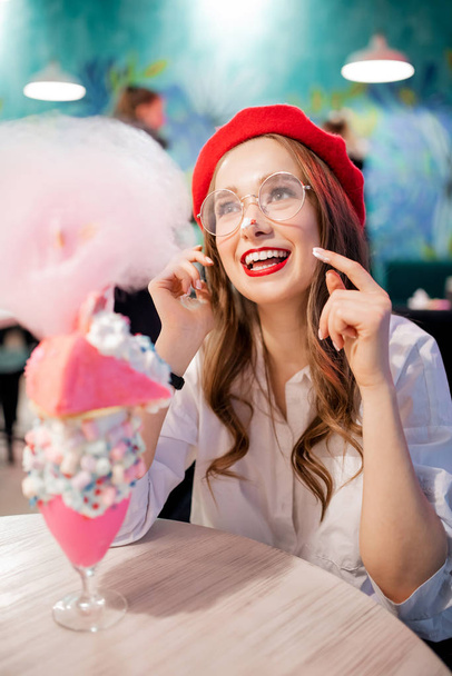 Francja słodki deser narodowy, dziewczyna w czerwonym berecie je różowe ciasto, nici cukierkowe i koktajl mleczny, śmieje się i uśmiecha - Zdjęcie, obraz