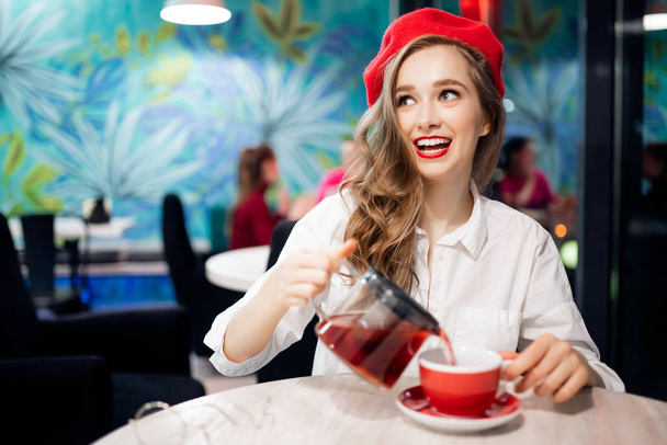 Romantisches Bild Frankreichs. Junges Mädchen in roter Baskenmütze mit welligem Haar lächelt und schenkt Tee zum Frühstück in Becher im Café ein - Foto, Bild