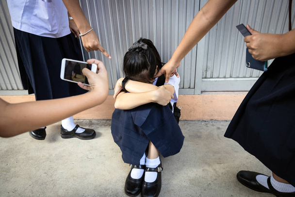 Mobbingprobleme in der Schule, traurig gestresste asiatische Schülerin weint auf dem Boden sitzend, Gruppe von Händen zeigt mit dem Finger auf verängstigte Schülerin, Mobbingopfer wird auf Handy aufgezeichnet - Foto, Bild