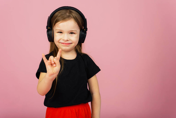 Een klein meisje in draadloze grote zwarte hoofdtelefoon op een roze pastelachtergrond kijkt naar de camera en toont een hoorns-up gebaar. Het concept van het luisteren naar rockmuziek. Plaats voor tekst, kopieerruimte - Foto, afbeelding