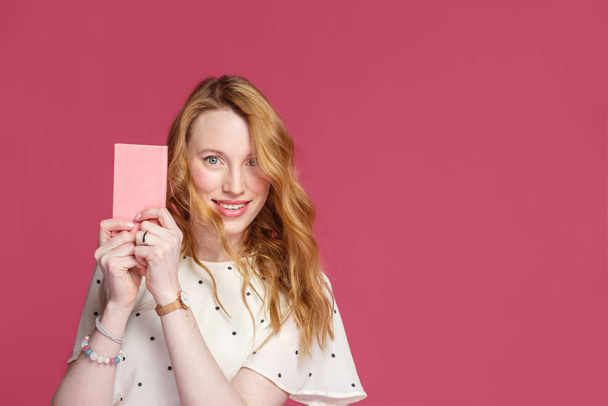 Веселая блондинка держит в руках розовую тетрадь, смотрит в камеру и улыбается, изолированная на розовом фоне
 - Фото, изображение