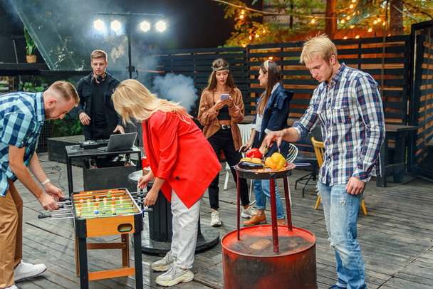 Close up of joyful guy with blond girl plays table football game while their male friend grilling vegetables in outdoors at weekend. Slow motion - Foto, Imagen
