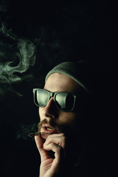 暗い背景に髭を生やした白いヒップスター喫煙者の肖像画。スタジオストックフォトタバコとマリファナの概念. - 写真・画像