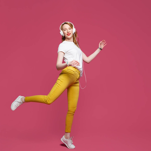 Beautiful blonde woman in white t-shirt, yellow trousers and white headphones listens to music on pink background. Dynamic studio photoshoot - Foto, immagini