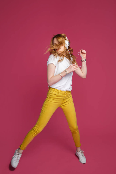 Beautiful blonde woman in white t-shirt, yellow trousers and white headphones listens to music on pink background. Dynamic studio photoshoot - Zdjęcie, obraz