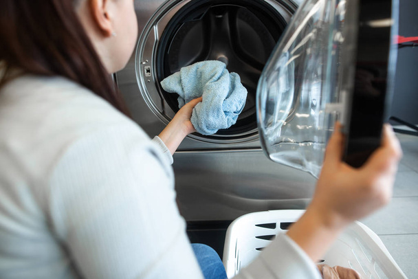 Çamaşırhanedeki çamaşır makinesine kirli çamaşırları koyan genç bir kadın. - Fotoğraf, Görsel