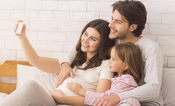 Schwangere Frau nimmt Selbstporträt mit Ehemann und süßer Tochter im Schlafzimmer - Foto, Bild