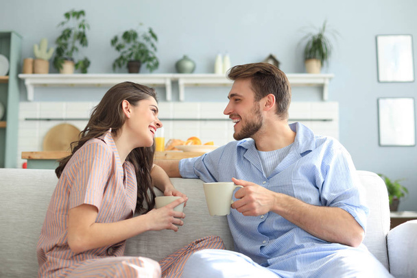 Beautiful young couple in pajamas is looking at each other and smiling on a sofa in the living room. - Foto, Imagen