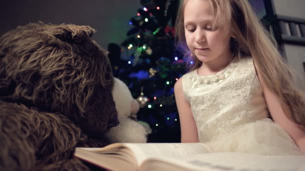 A little blonde girl in a festive dress with a book in her hands sits next to soft toys against the background of a Christmas tree and reads a book leading the page with her place. - Materiaali, video