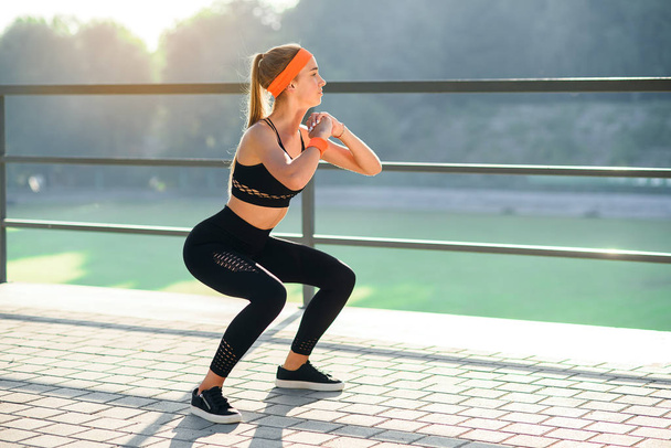 Gorgeous sporty girl in black sportswear and orange headband which squating during fitness training at stadium outdoors. - Photo, Image