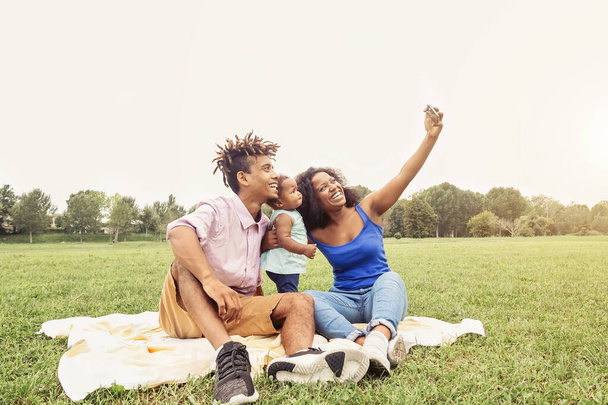 Happy African family doing selfie photo with mobile phone in a public park outdoor - Mother and father having fun with their daughter during a weekend sunny day - Love and happiness concept - 写真・画像