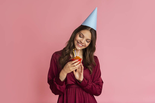 Młoda dziewczyna w kapeluszu urodzinowym patrząc na babeczkę z zapalonymi świeczkami - Zdjęcie, obraz