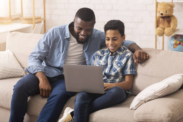 Отец учит сына пользоваться ноутбуком, сидя дома на диване
 - Фото, изображение