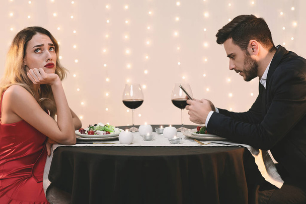 Unhappy woman sitting alone while her boyfriend stuck into his phone - 写真・画像
