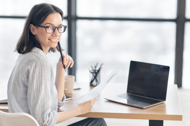 Усміхнена молода жінка-працівник сидить з паперами і ноутбуком
 - Фото, зображення