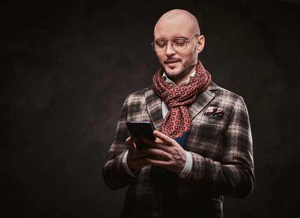 Erfolgreicher Geschäftsmann posiert mit karierter Jacke, Brille und Schal im Studio und hält ein Smartphone in der Hand - Foto, Bild