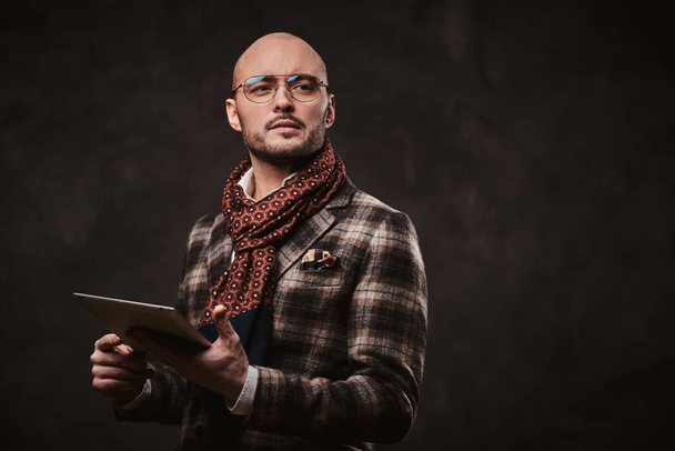 Successful stylish businessman posing in a studio wearing checkered jacket, glasses and scarf while holding a tablet - 写真・画像