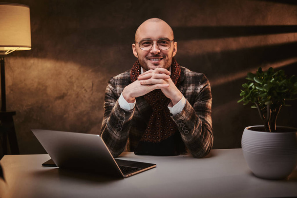 Bald europeu bem vestido sorridente empresário sentado no escritório em uma mesa com notebook, vestindo jaqueta elegante e um lenço
 - Foto, Imagem