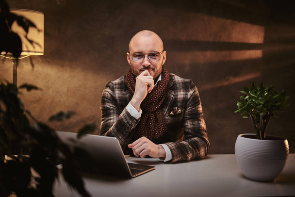 Лысый европейский хорошо одетый серьезный бизнесмен сидит в офисе за столом с ноутбуком, одетый в стильную куртку и шарф
 - Фото, изображение