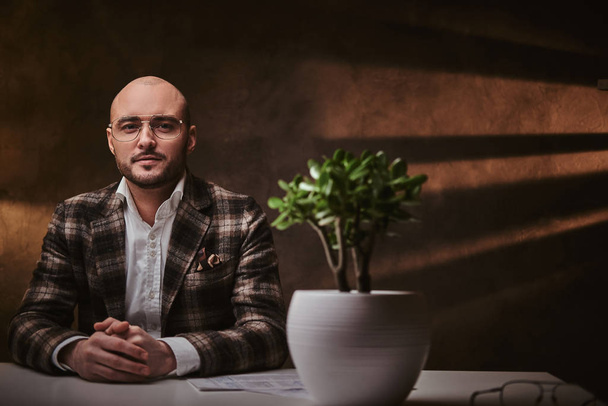 Καραφλός Ευρωπαίος καλοντυμένος επιχειρηματίας που κάθεται στο γραφείο σε ένα τραπέζι με σημειωματάριο, φορώντας κομψό σακάκι και γυαλιά - Φωτογραφία, εικόνα