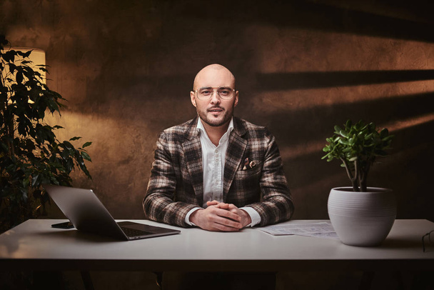 Καραφλός Ευρωπαίος καλοντυμένος επιχειρηματίας κάθεται στο γραφείο σε ένα τραπέζι με σημειωματάριο, φορώντας κομψό σακάκι - Φωτογραφία, εικόνα