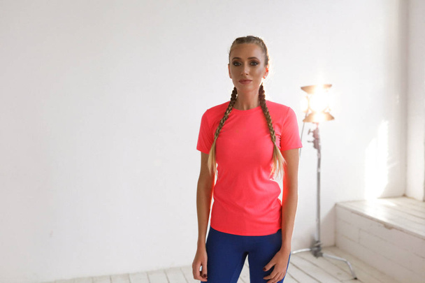Νεαρό ξανθό κορίτσι με καλλίγραμμο σώμα στέκεται σε εσωτερικούς χώρους κοντά στο λευκό τοίχο και φωτεινό φως. Φορώντας ροζ και μπλε άνετα αθλητικά ρούχα, κομψά μαλλιά και λαμπερό μακιγιάζ.   - Φωτογραφία, εικόνα