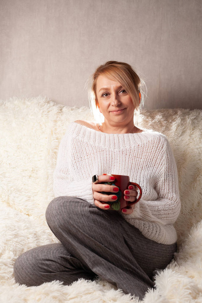 Πορτρέτο μιας ενήλικης γυναίκας σε ένα άνετο δωμάτιο στον καναπέ με ένα φλιτζάνι τσάι / καφέ. - Φωτογραφία, εικόνα