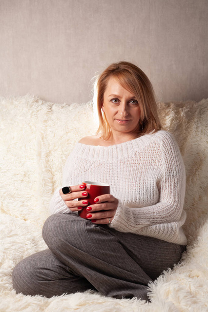Retrato de uma mulher adulta em um quarto acolhedor no sofá com uma xícara de chá / café
. - Foto, Imagem