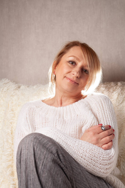 A beautiful adult woman is sitting in a room on a sofa. Portrait of a blonde at home. - Foto, Bild