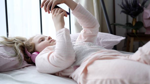 moderni elämä sukupolven Z teini tyttö pyjamassa ja kuulokkeet huoneessa sängyllä kuuntelee musiikkia älypuhelimesta
. - Valokuva, kuva