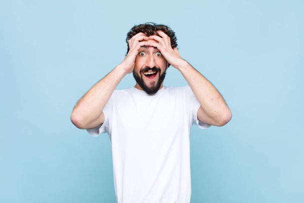 young crazy bearded man raising hands to head, open-mouthed, feeling extremely lucky, surprised, excited and happy against flat wall - Foto, imagen