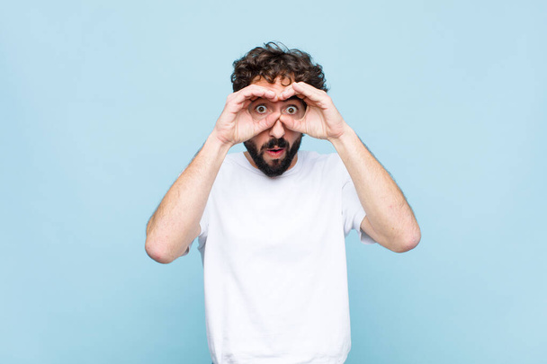 young crazy bearded man feeling shocked, amazed and surprised, holding glasses with astonished, disbelieving look against flat wall - Fotoğraf, Görsel