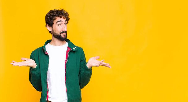 young crazy bearded man feeling puzzled and confused, doubting, weighting or choosing different options with funny expression against flat wall - Foto, imagen